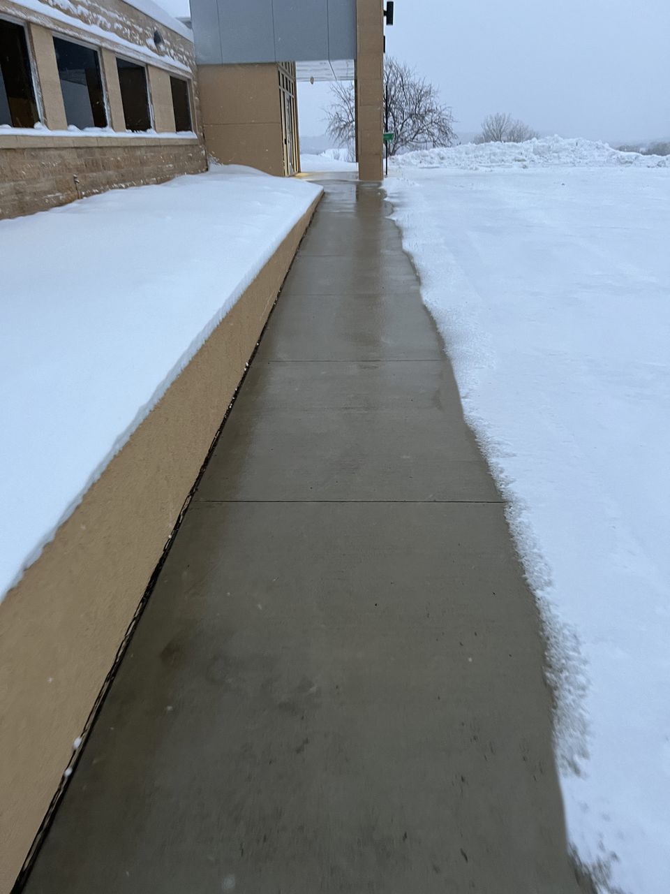 Clear concrete walkway beside a building after a snowfall, with snow covering the ground on both sides